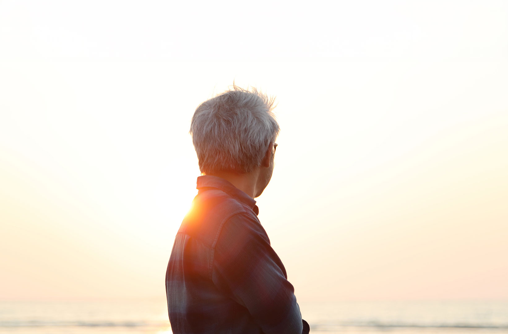 Grey-haired older person seen from behind in front of a bright, hazy sunset over the sea